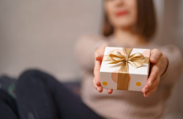 Mãos oferecem caixa de presente branca com laço dourado e bolinhas coloridas.