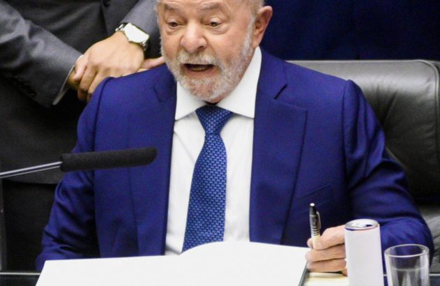 Lula, de terno azul, sentado à mesa, escreve em um livro aberto com uma caneta. Ele tem a boca aberta.