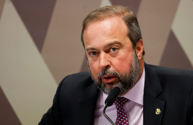 Homem de terno e barba discursa em dois microfones. Ambiente oficial.