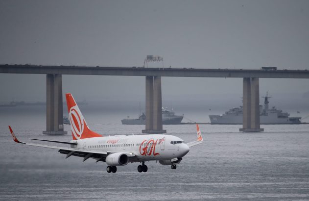 Avião da GOL pousando sobre a água com a Ponte Rio-Niterói e navios militares ao fundo.