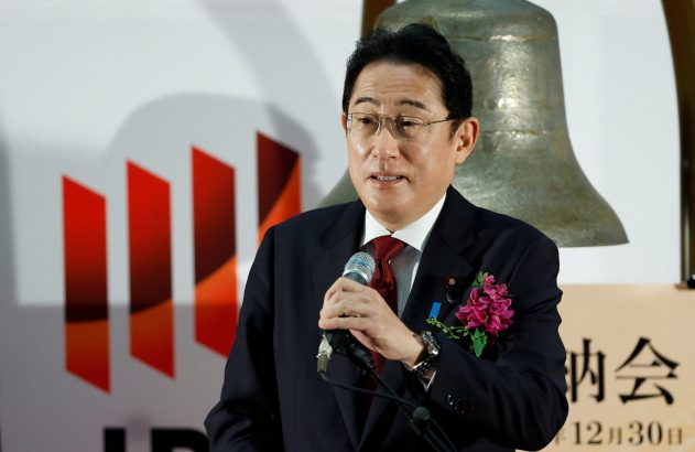 Homem de terno e óculos discursando com microfone. Ao fundo, sino grande e logotipo vermelho.