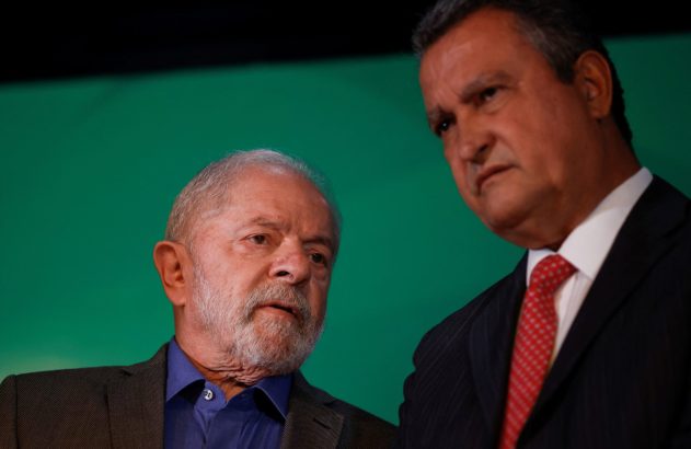 Lula, com barba branca, e Rui Costa, de terno e gravata vermelha, observam atentos em frente a um fundo verde.