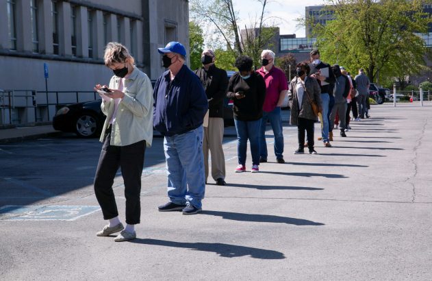 Longa fila de pessoas usando máscaras em um estacionamento ao ar livre. Muitos olham seus celulares.