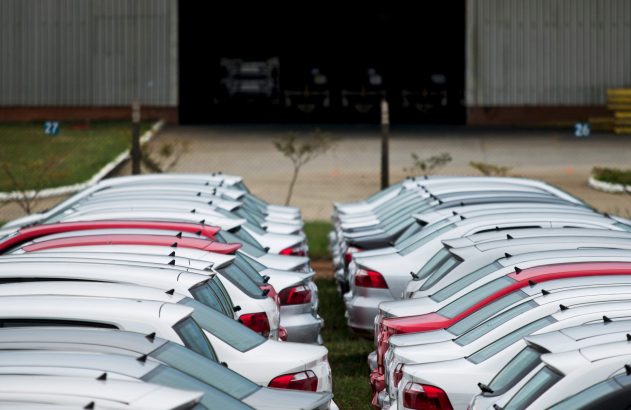 Fileiras de carros novos, brancos e vermelhos, estacionados em pátio de fábrica. Grande galpão industrial ao fundo.
