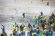 Multidão com bandeiras do Brasil e barreiras metálicas caídas, em meio a fumaça.