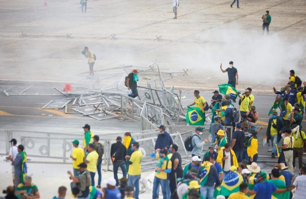 Multidão com bandeiras do Brasil e barreiras metálicas caídas, em meio a fumaça.