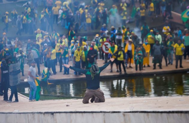 Homem ajoelhado com braços abertos; ao fundo, multidão em amarelo e verde, fumaça e pessoa com escudo.