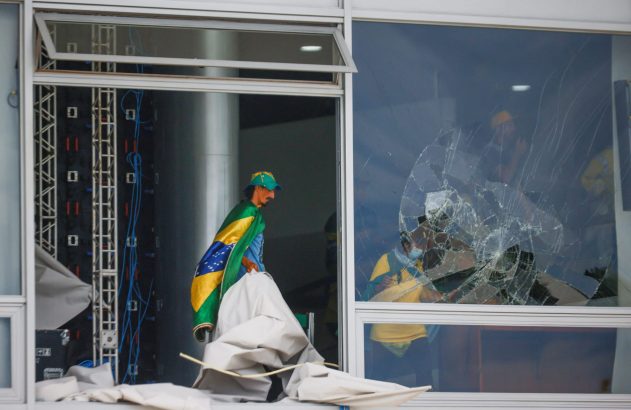Manifestante com bandeira do Brasil na entrada de prédio danificado, vidro estilhaçado mostra mais pessoas.