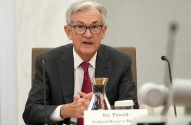 Jay Powell, de terno, gravata e óculos, sentado à mesa, falando. Letreiro 'Jay Powell Federal Reserve Bo' visível.