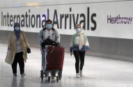 Três passageiros de máscara chegam ao Aeroporto de Heathrow. Um empurra bagagem. Sinal 'International Arrivals'.