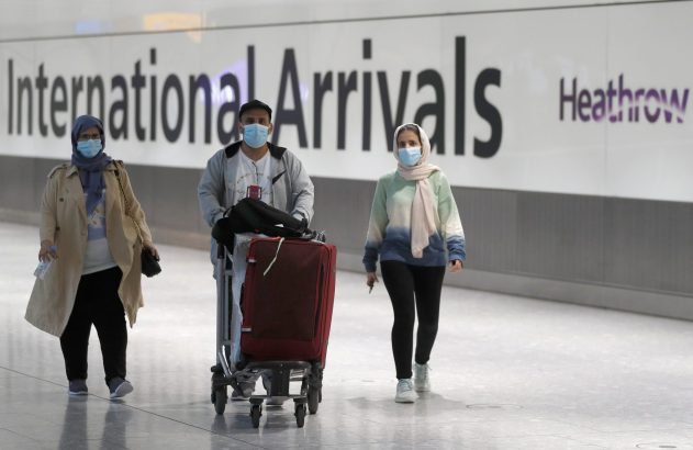 Três passageiros de máscara chegam ao Aeroporto de Heathrow. Um empurra bagagem. Sinal 'International Arrivals'.