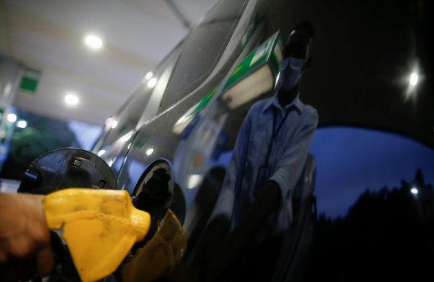 Mão abastecendo carro com bico amarelo. Reflexo de pessoa mascarada no veículo.