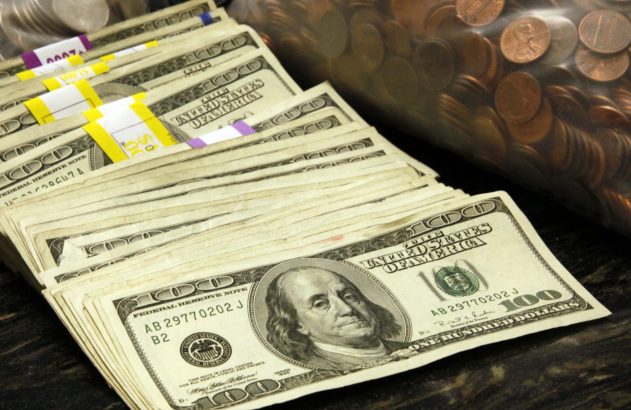 Pilhas de notas de US$100 com Benjamin Franklin e sacos de moedas, incluindo centavos, sobre uma superfície escura.