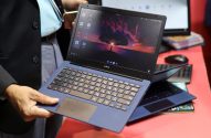 Pessoa segura laptop azul aberto com tela ligada mostrando a área de trabalho. O nome 'JioBook' é visível.