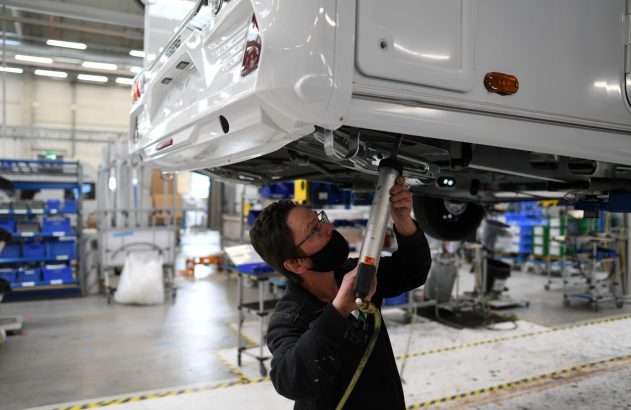 Pessoa com máscara aplica selante sob a parte traseira de um motorhome branco. Cena em fábrica de montagem veicular.