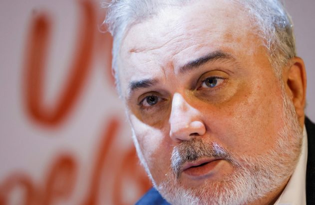 Close-up do rosto de um homem com barba e cabelo grisalhos. Ele olha para o lado, boca entreaberta. Fundo desfocado.