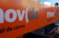 Letreiro laranja "Movida aluguel de carros" em vitrine. Pessoa ao fundo.