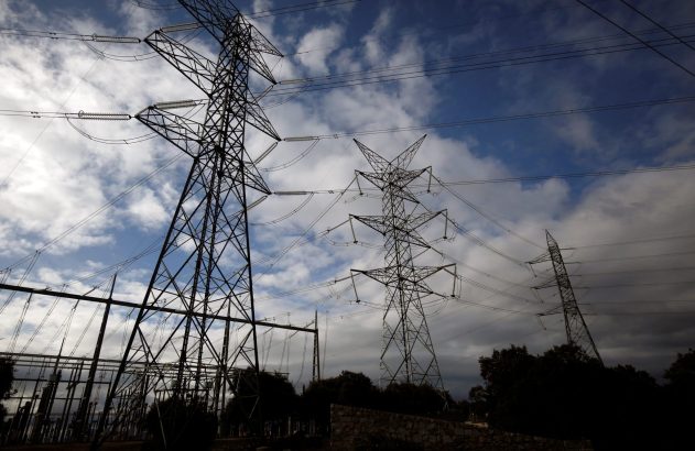 Torres de alta tensão e fios elétricos em silhueta sob céu nublado. Uma subestação e vegetação escura em primeiro plano.