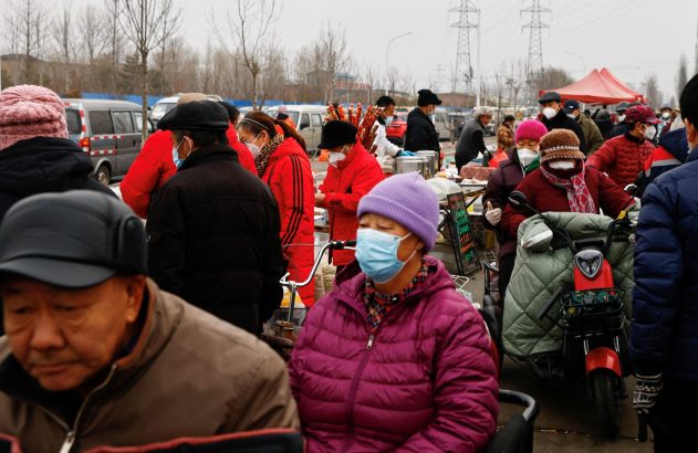 Mercado de rua movimentado com pessoas usando máscaras. Barracas, veículos e mercadorias são visíveis.