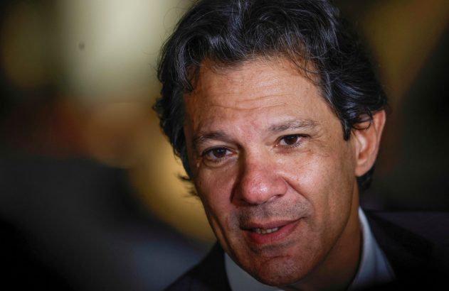 Close-up de um homem com cabelo escuro, olhar direto e sorriso sutil. Fundo escuro e desfocado.