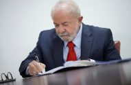 Lula, de terno e gravata vermelha, escreve em documento à mesa. Óculos à esquerda.