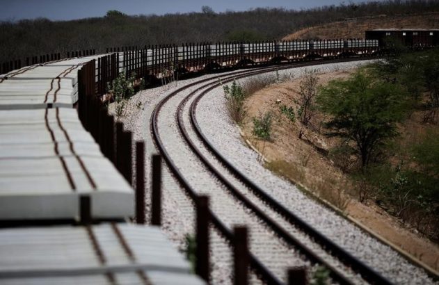 Trilhos de trem curvados com um trem transportando dormentes ao longe. Pilhas de dormentes novos à esquerda.