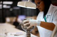 Pessoa de luvas e jaleco examina vaso metálico ornamentado sob luz de lupa em um laboratório.