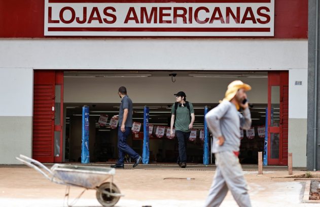 Entrada de Lojas Americanas com pessoas caminhando. Um carrinho de mão e um homem ao telefone em primeiro plano.