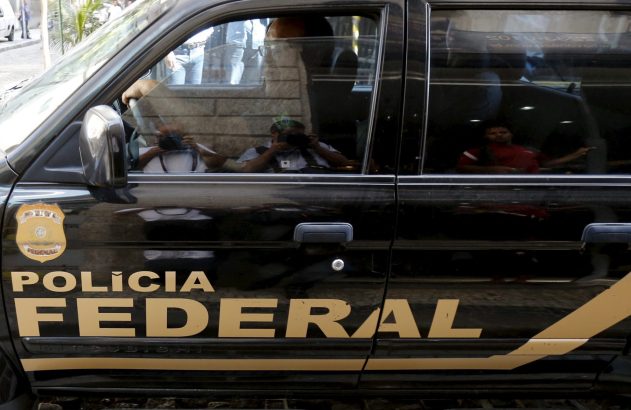 Viatura preta da Polícia Federal com brasão e texto em dourado. Reflexos de pessoas e fotógrafos nas janelas.