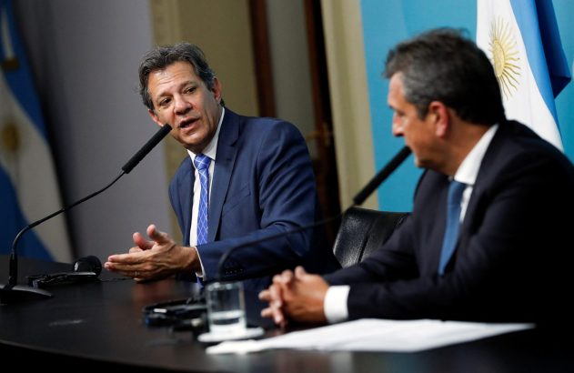 Homem em terno fala ao microfone em mesa com outro homem; bandeiras da Argentina ao fundo.