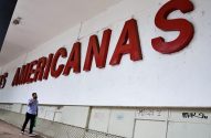 Homem caminha sob grande letreiro vermelho "AMERICANAS" em parede branca, olhando para cima.
