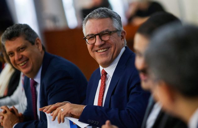 Homem de terno azul e óculos sorri para a câmera em reunião. Outro homem sorridente está ao seu lado.