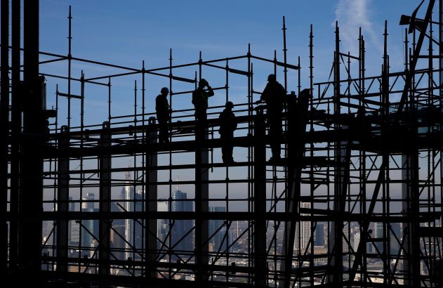 Silhuetas de operários em andaimes de construção. Céu azul e paisagem urbana de prédios ao fundo.