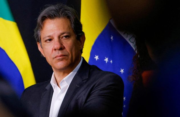 Homem com cabelos grisalhos, camisa branca e terno escuro, olha à frente. Bandeiras do Brasil ao fundo.