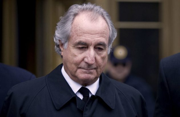 Homem de cabelos grisalhos em casaco escuro e gravata preta, olhando para baixo em ambiente externo.