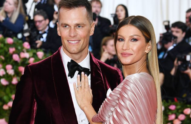 Tom Brady e Gisele Bündchen sorrindo. Gisele, de vestido rosa, toca o terno bordô de Tom.