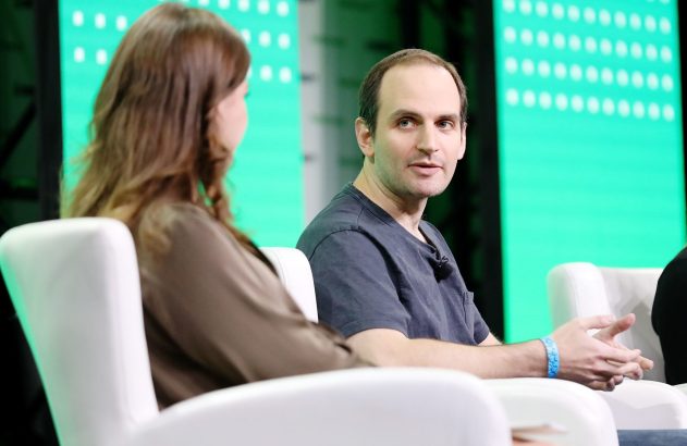 Homem de camiseta escura e mulher de costas sentados em cadeiras brancas, em evento com telas verdes. O homem olha para frente.