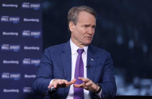 Homem de terno azul e gravata roxa gesticula, com logotipos "Bloomberg" e "Wall Street" ao fundo.