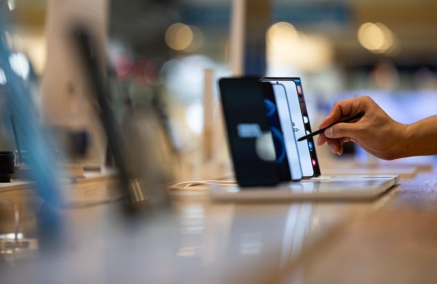 Mão usando uma caneta stylus em um smartphone dobrável com a tela aberta, em exposição em uma loja.