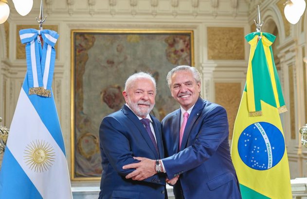 Lula e Alberto Fernández sorriem em encontro, com bandeiras do Brasil e Argentina ao fundo.