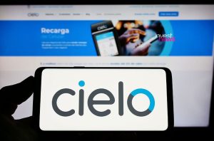 Mão segura celular com logo Cielo. Monitor exibe website da Cielo com ‘Recarga’ e ‘Invest News’.