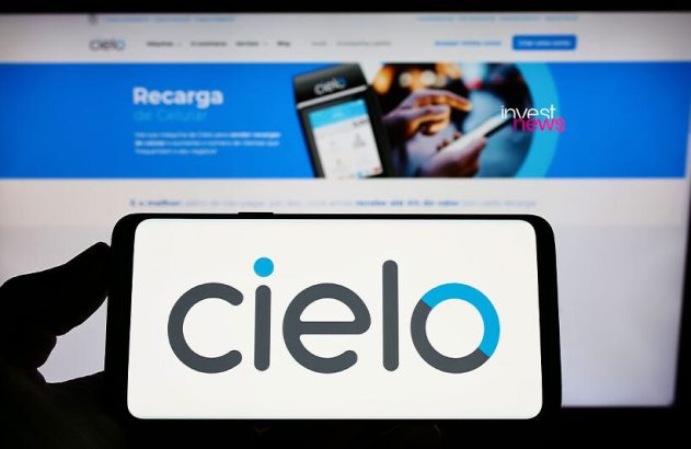 Mão segura celular com logo Cielo. Monitor exibe website da Cielo com ‘Recarga’ e ‘Invest News’.