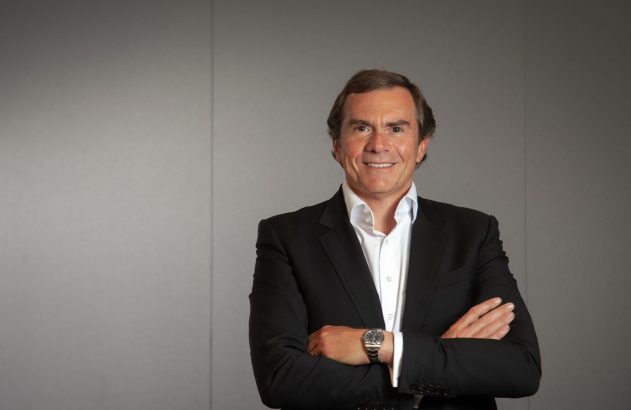 Homem de terno preto sorri com braços cruzados contra parede cinza.