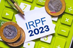Nota com "IRPF 2023", caneta, moedas de Real e calculadora verde.