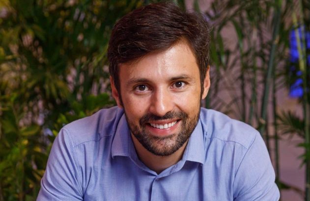 Retrato de homem sorrindo, com barba e camisa azul, olhando para a câmera. Fundo verde.