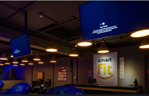 Interior de academia com TVs mostrando 'Sem Sinal', logo Smart Fit, iluminação quente e equipamentos.