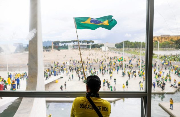 Homem de costas em camisa amarela 'Deus Acima de Tudo' segura bandeira do Brasil, olhando multidão por janela rachada.