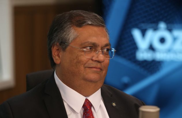 Homem de terno, óculos, com leve sorriso, olhando para a direita. Fundo azul desfocado com o logo 'Voz do Brasil'.