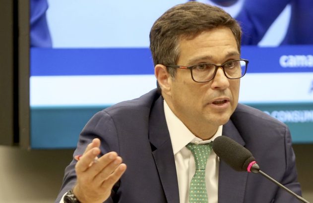 Homem de terno, óculos e gravata verde fala com microfone, gesticulando. Tela ao fundo com textos "camara" e "consumidor".