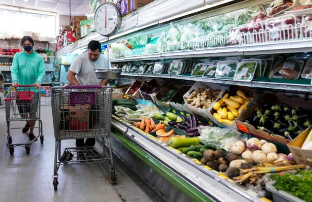 Duas pessoas em mercado: uma empurra carrinho, outra pesa vegetais frescos no balcão de hortifrúti.